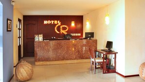 Zona de recepción de hotel con mostrador de mármol, escritorio con computadora, una silla y un jarrón decorativo grande.