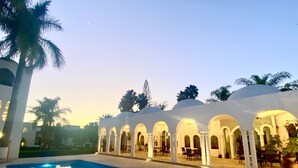 A pool area with a clear reflection, surrounded by white columns and arches, palm trees, and a building with a dome-shaped roof. - Aldea del Bazar (Tehuacán)