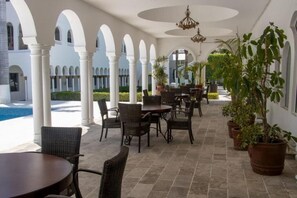 Un espace extérieur abrité, aménagé avec des tables et des chaises, entouré de colonnes blanches et de plantes en pot.