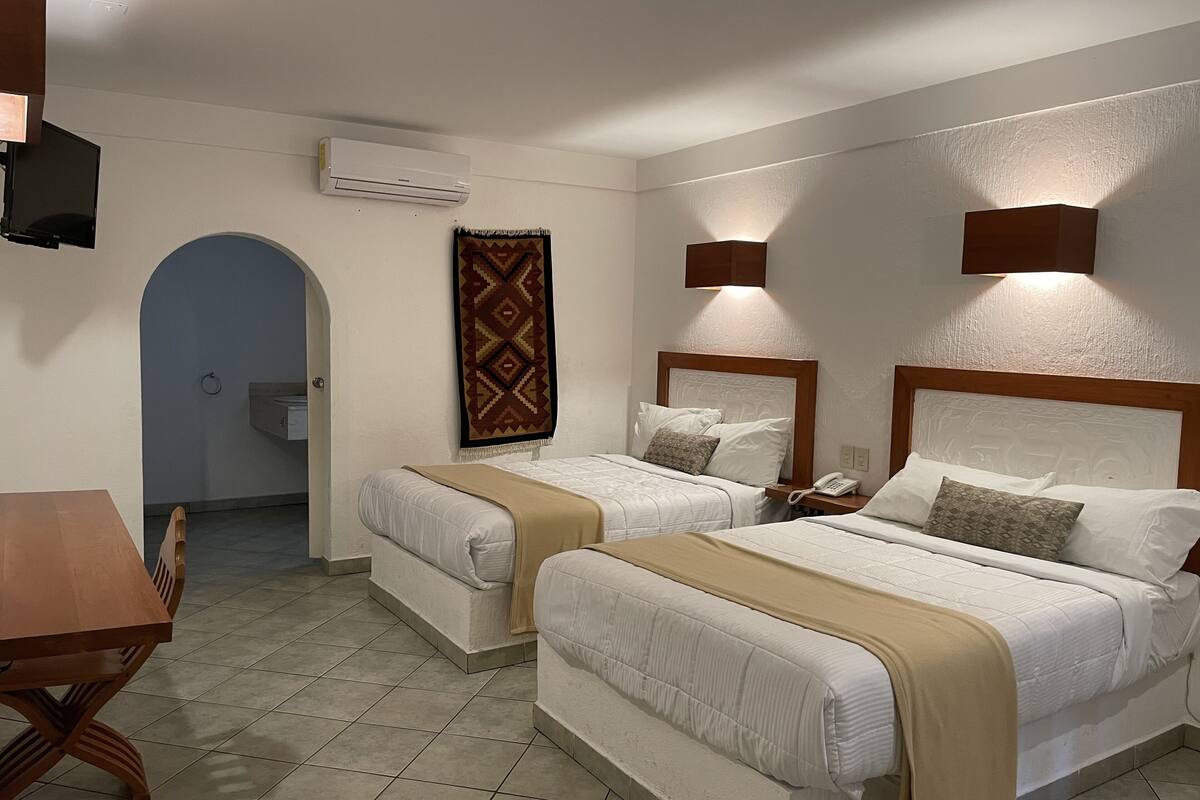 A hotel room with two beds, a wooden table, an air conditioning unit, a patterned rug, and a wall-mounted phone.
