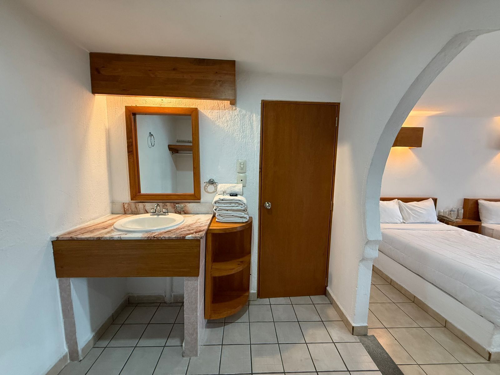 Habitación de hotel con tocador de madera, lavamanos, espejo, puerta y cama con ropa blanca.
