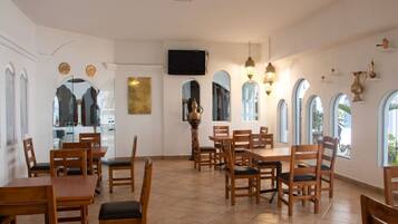 A dining area with wooden tables and chairs, a television mounted on the wall, and arched windows.