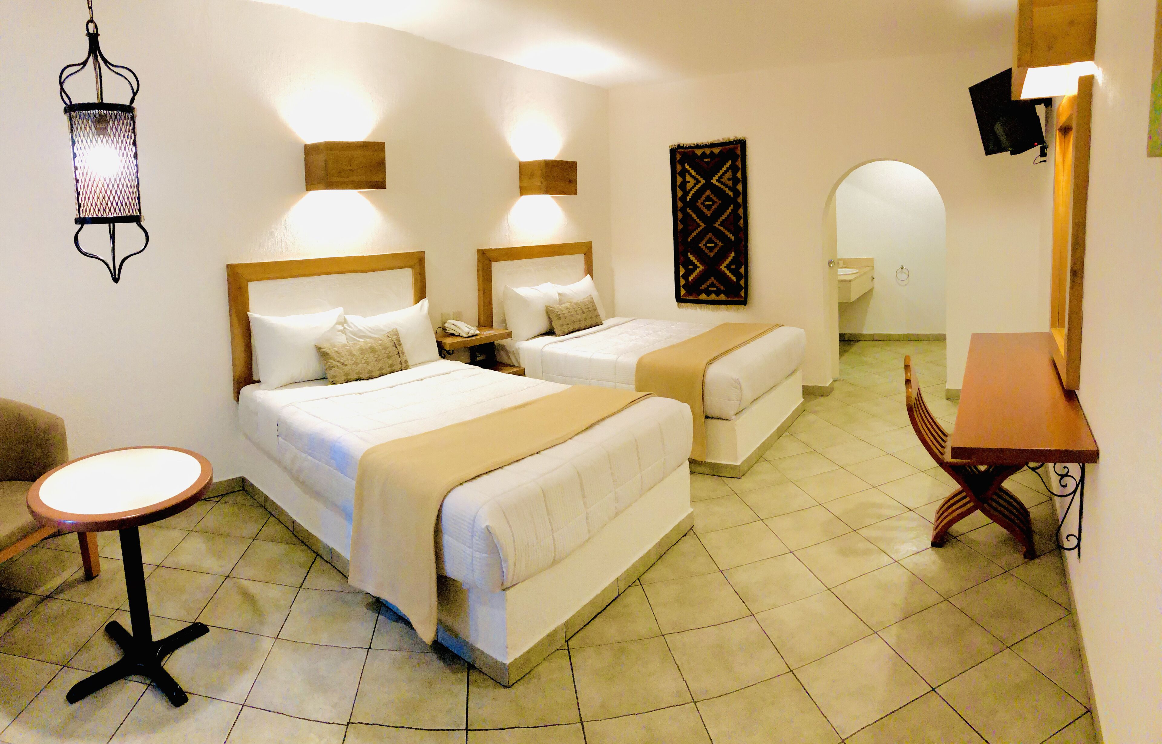 Habitación de hotel con dos camas, una mesita redonda pequeña, una silla, televisión y baño visible a través de un arco.