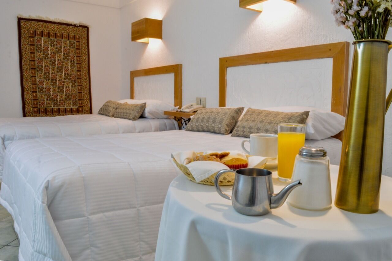 A hotel room with two beds, a breakfast table with a basket of pastries, a coffee pot, a teapot, a glass of orange juice, and a vase with flowers.