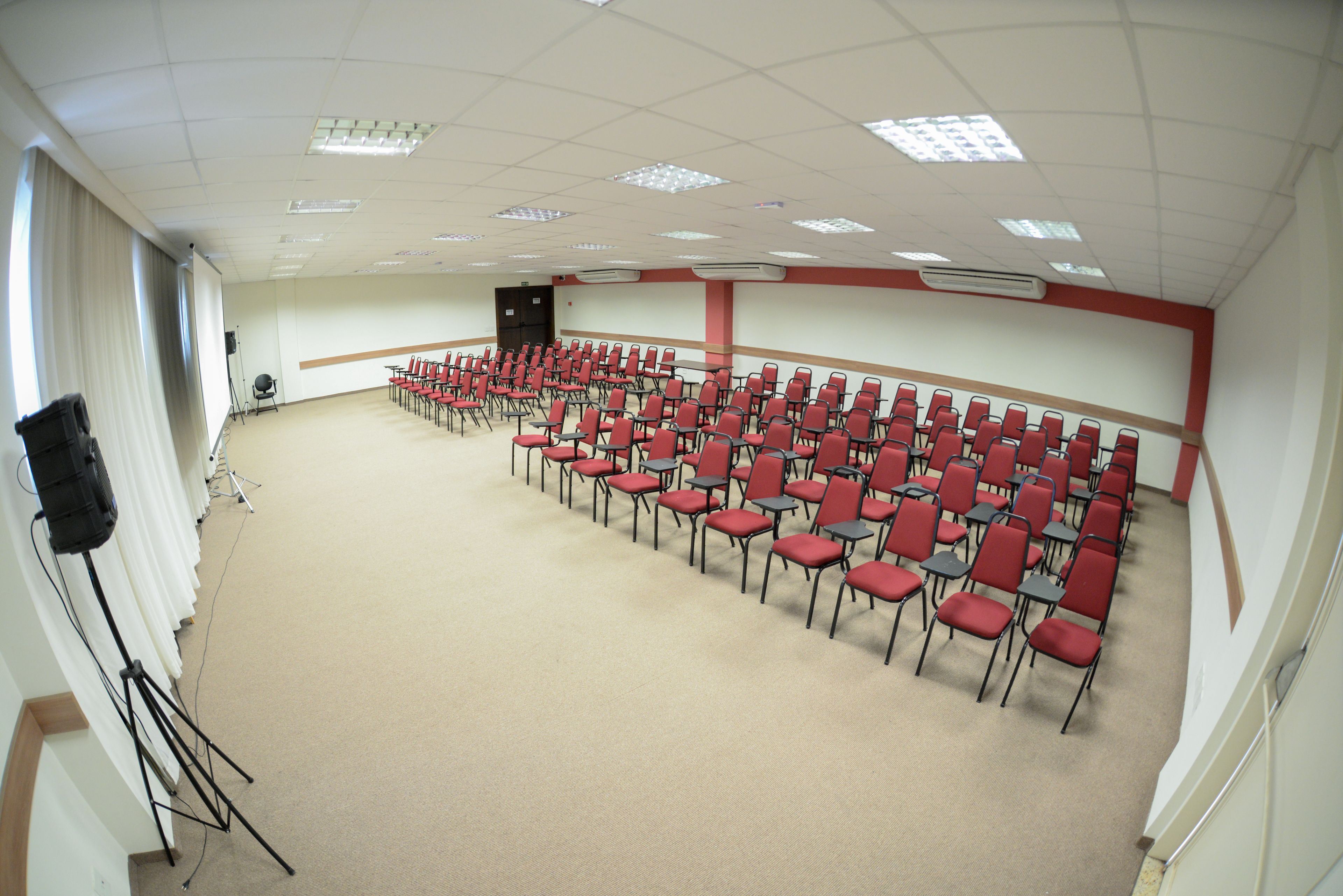 A conference room with rows of red chairs, a stage with a speaker, and ceiling lights.
