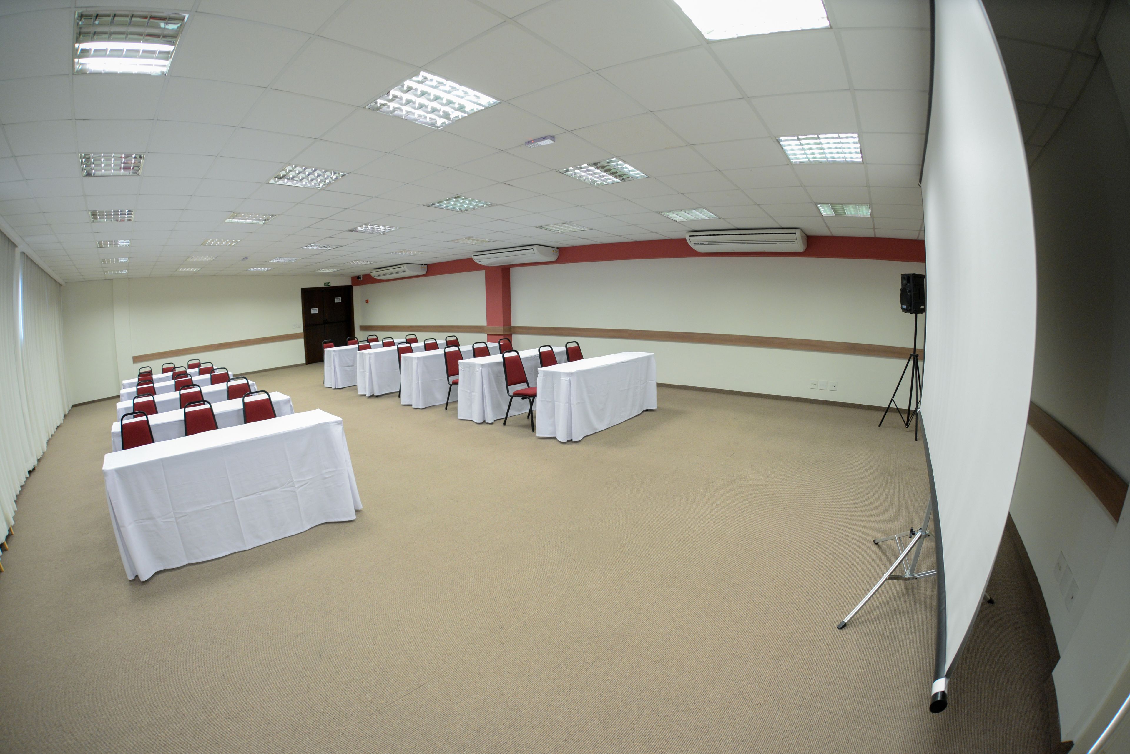 Une salle de conférence avec des rangées de tables et de chaises, des unités de climatisation et un trépied posé sur le sol.