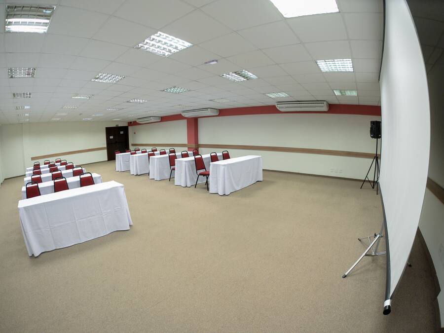 Une salle de conférence avec des rangées de tables et de chaises, des unités de climatisation et un trépied posé sur le sol.