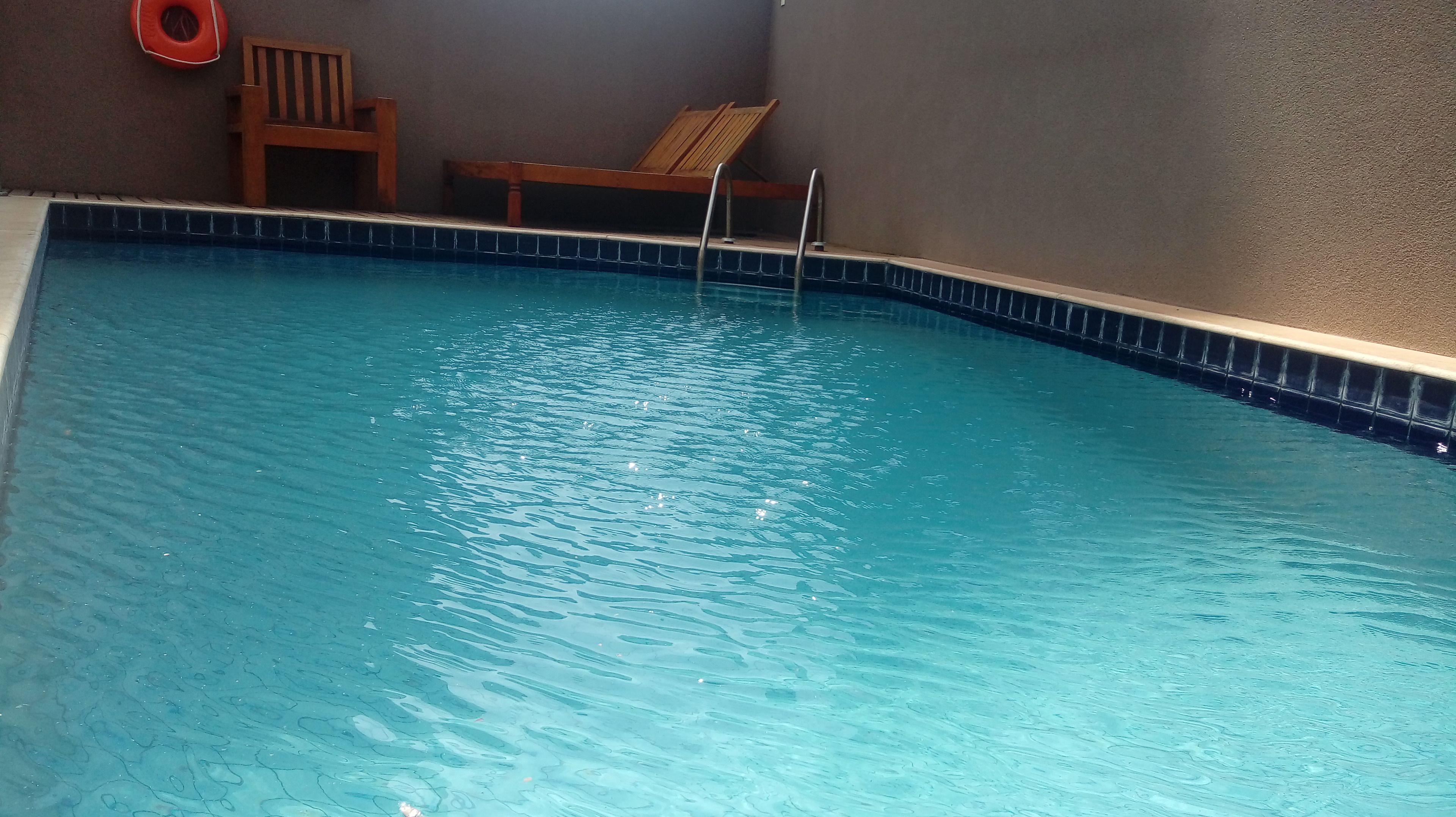 A swimming pool with clear water, wooden lounge chairs, and a lifebuoy on the wall.