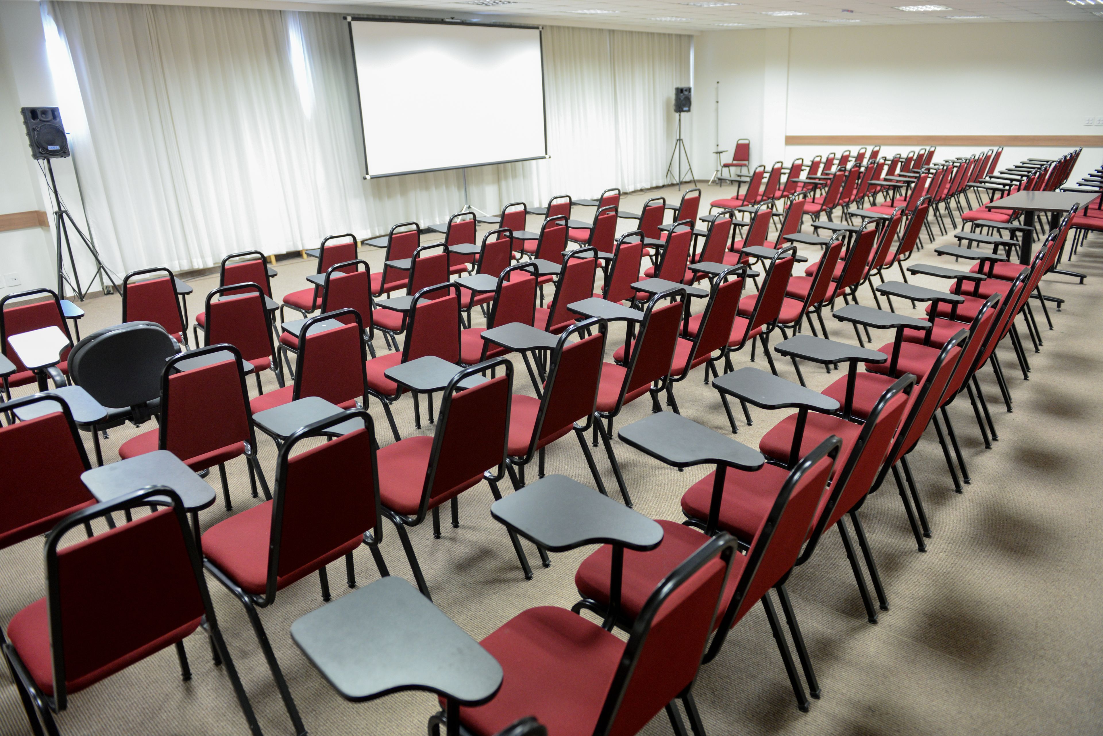 Uma sala de conferências com fileiras de cadeiras vermelhas e mesas pretas, uma tela de projetor e um orador em um suporte.