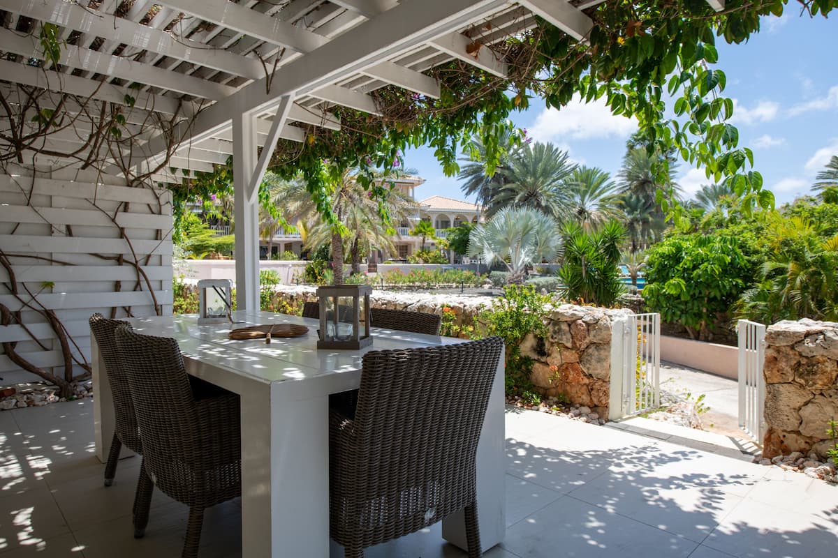 Un patio exterior con una pérgola blanca, sillas de mimbre y una mesa con un farol.