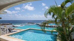 A pool area with clear blue water, a view of the ocean, and a white fence with lounge chairs. - Curacao Luxury Holiday Rentals (Willemstad)