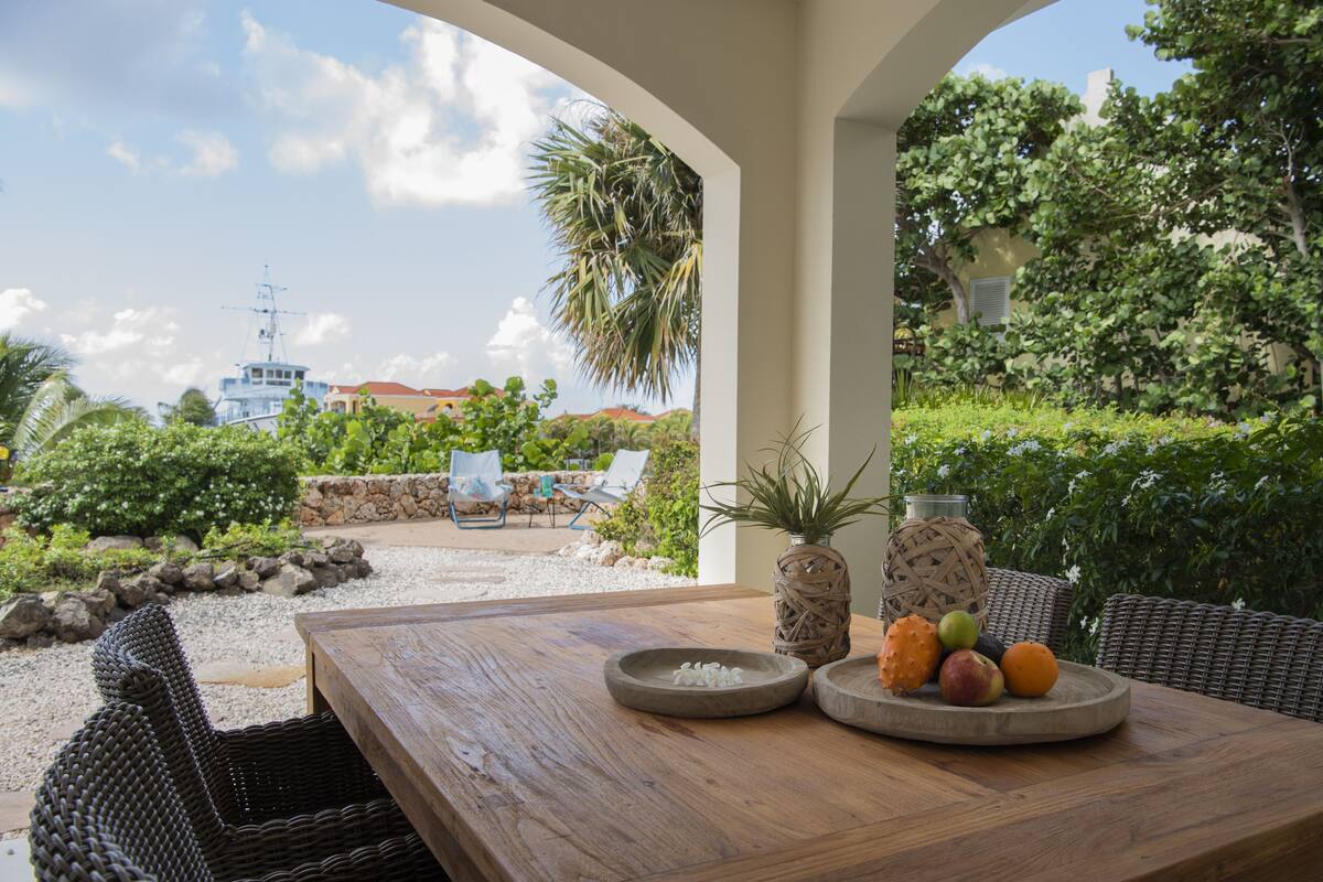 Una mesa de madera con un frutero y una cesta tejida, ubicada en un espacio exterior cubierto con mobiliario de mimbre y vistas a un edificio y vegetación al fondo.