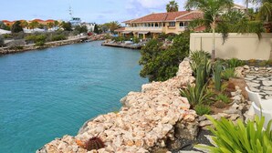 A coastal view with a marina, a stone pathway, and a building with a red-tiled roof. - Curacao Luxury Holiday Rentals (Willemstad)