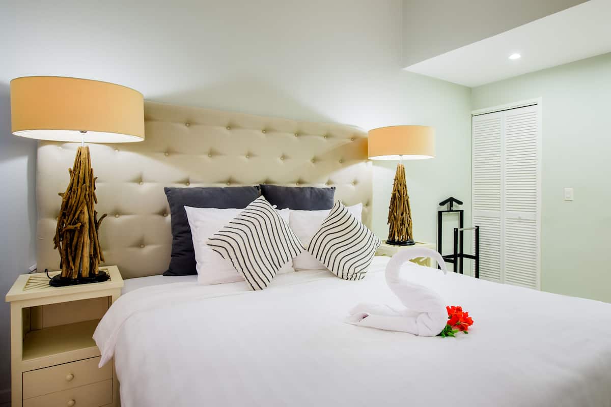 Una cama bien hecha con almohadillas a rayas, un edredón blanco y una toalla con flores rojas. Hay dos lámparas de noche con pantallas color beige y una escultura de madera decorativa en la mesita de noche.