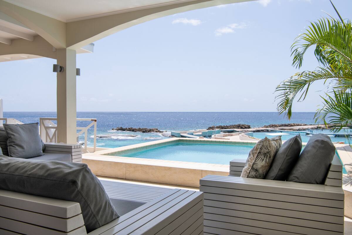 Zona de piscina con vista al océano, un área de descanso con cojines y una palmera.