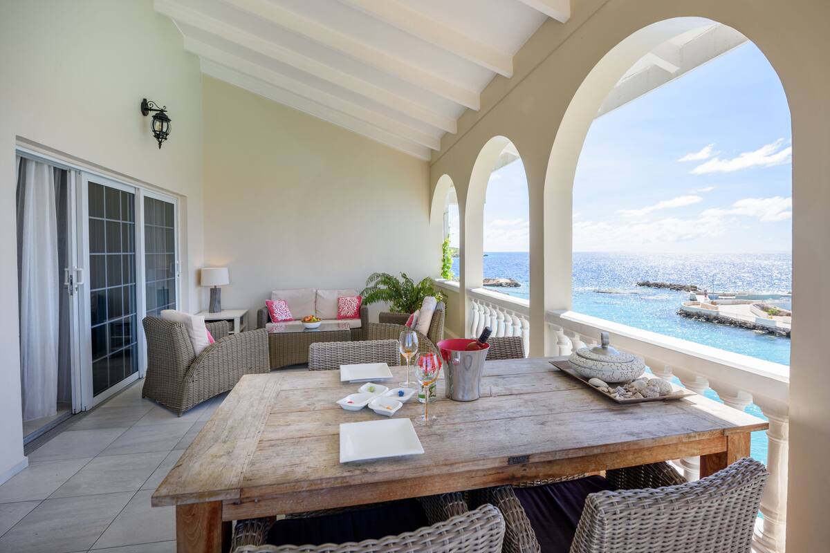 Un amplio balcón con mesa de madera dispuesta para comer, mobiliario de mimbre y vista al océano.
