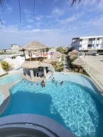 Una piscina con una estructura de techo de paja, una zona de estar cubierta y un edificio al fondo.
