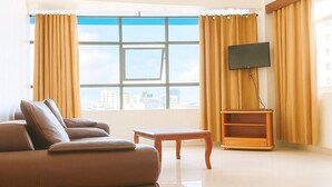 A spacious living room with a large window, a ceiling fan, a flat-screen TV, and a wooden coffee table. - Rainbow Hotel (Dar es Salaam)