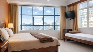 A hotel room with a large bed, a bedside table with a lamp, a TV mounted on the wall, a ceiling fan, and a window with a city view. - Rainbow Hotel (Dar es Salaam)