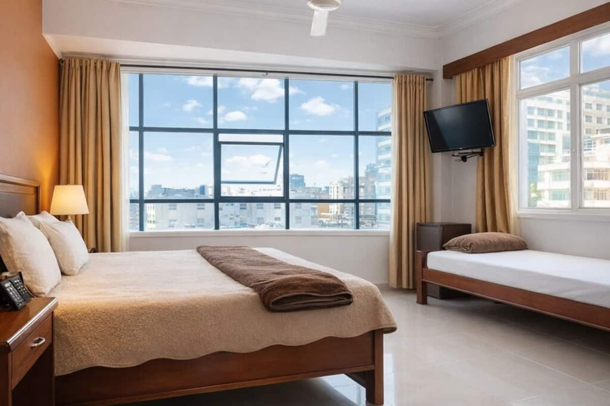 Habitación de hotel con una cama grande, una mesita de noche con lámpara, televisor montado en la pared, ventilador de techo y ventana con vista a la ciudad.
