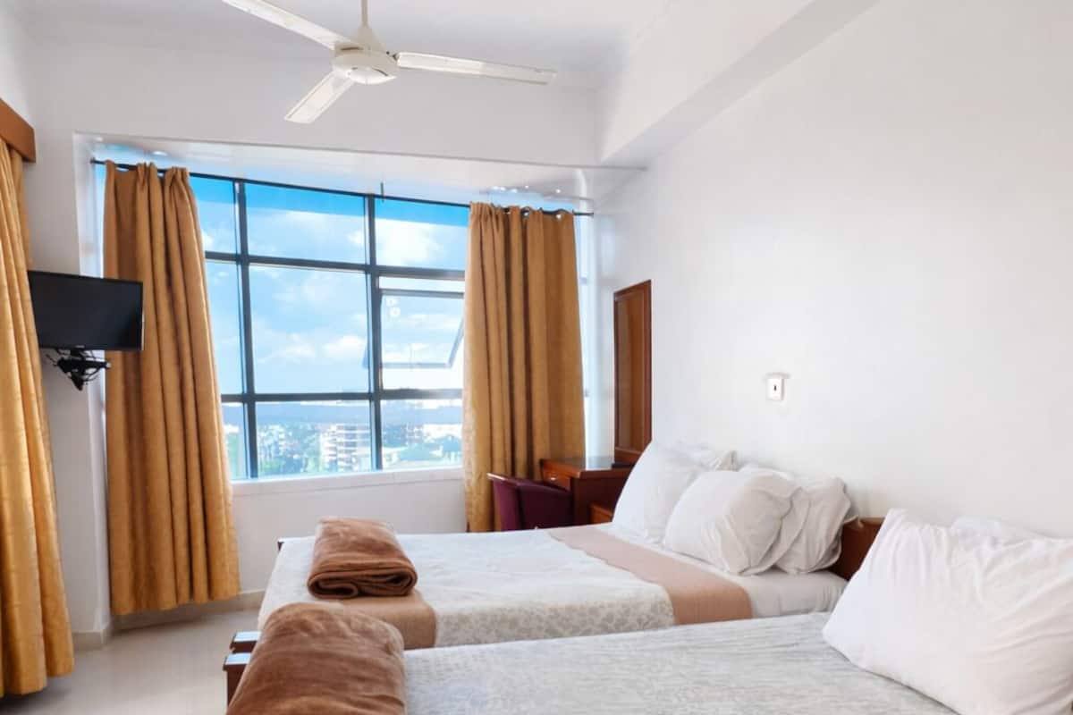 Habitación de hotel con dos camas, ventilador de techo, televisión y un ventanal grande con cortinas.