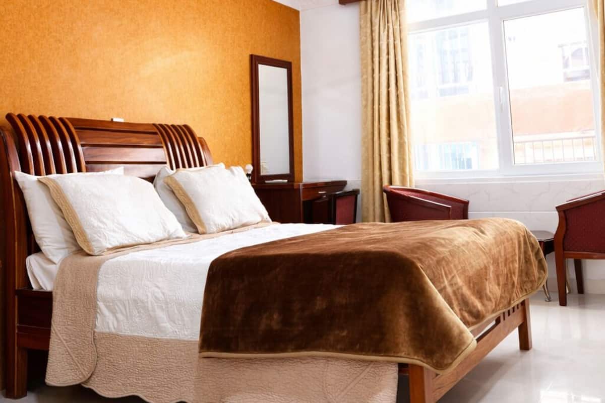 Una habitación de hotel con una cama grande, un ventilador de techo, una ventana con cortinas y un cabecero de madera.