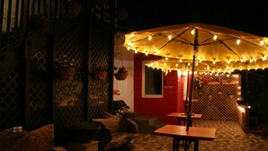 An outdoor patio area with wooden tables and chairs, a large umbrella with string lights, and potted plants. - The Loft at Menorcan Square (St. Augustine)