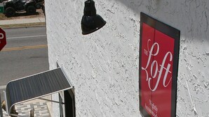A building with a sign reading "The Loft Nightly Accommodations" and a potted plant with pink flowers at the entrance. - The Loft at Menorcan Square (St. Augustine)