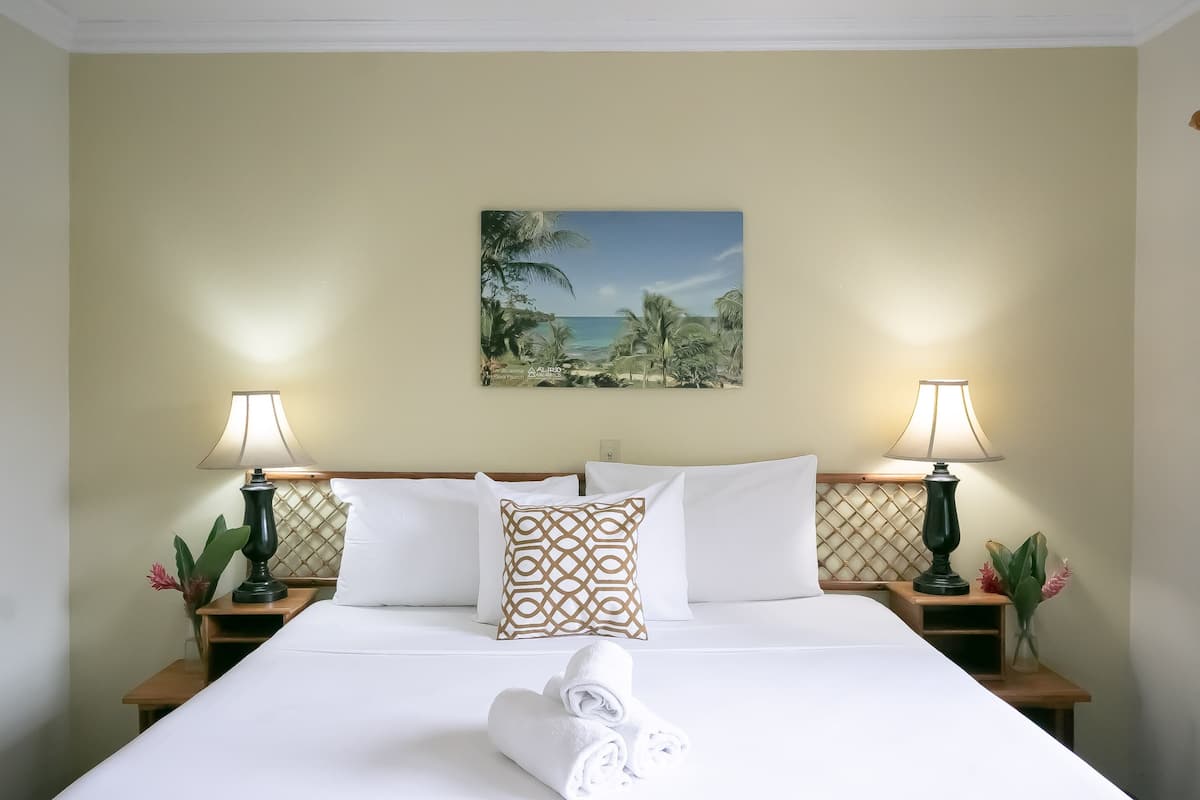 A neatly made bed with white linens, two bedside lamps, a decorative pillow, and a painting of a beach scene on the wall.