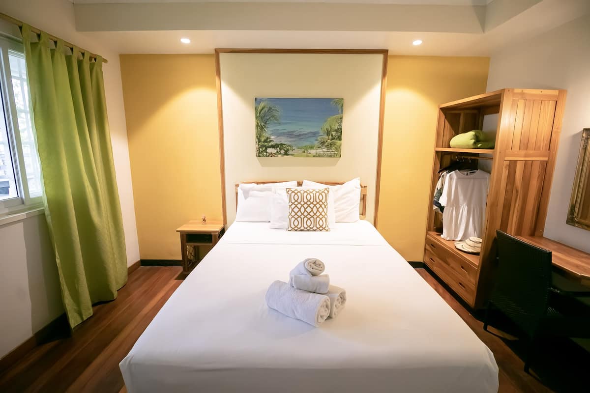 A neatly made bed with a towel, a wooden wardrobe, and a painting of a tropical scene on the wall.