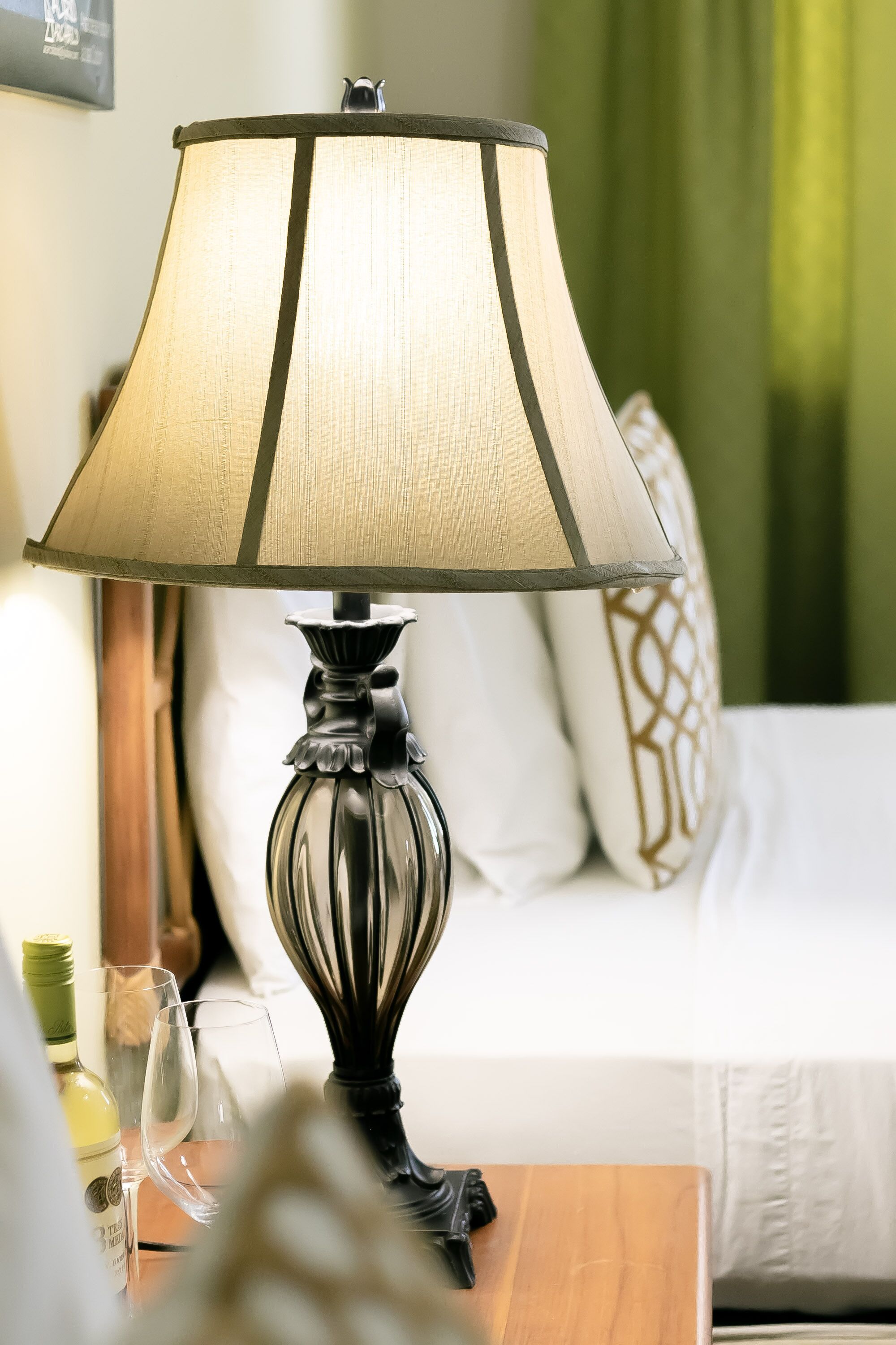 Une lampe dotée d’un abat-jour beige et d’un pied en bois, placée sur une table en bois dans une pièce où se trouvent un lit et, en arrière-plan, un rideau vert.