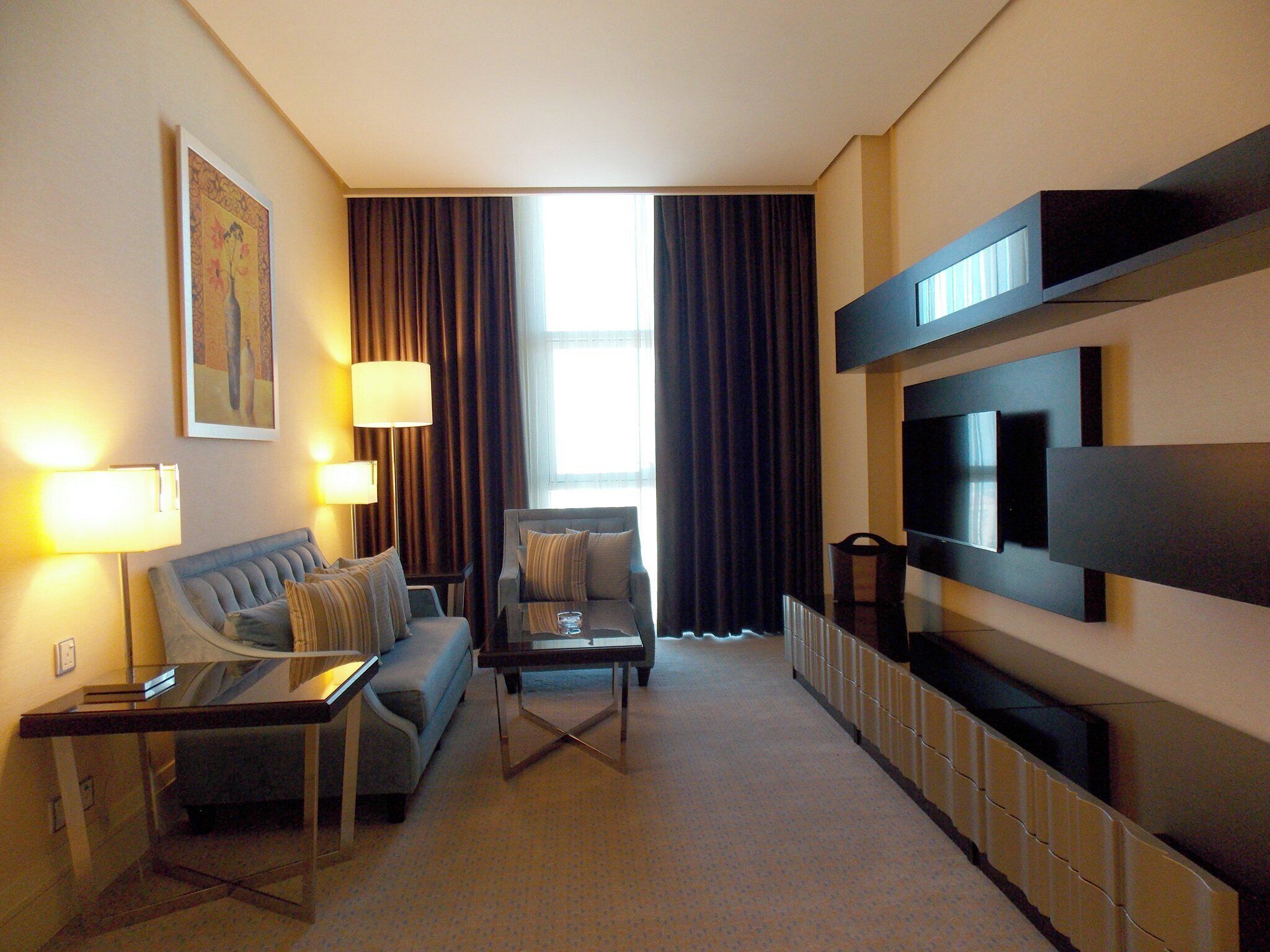 A modern hotel room with a sofa, armchair, coffee table, and a flat-screen TV mounted on the wall.