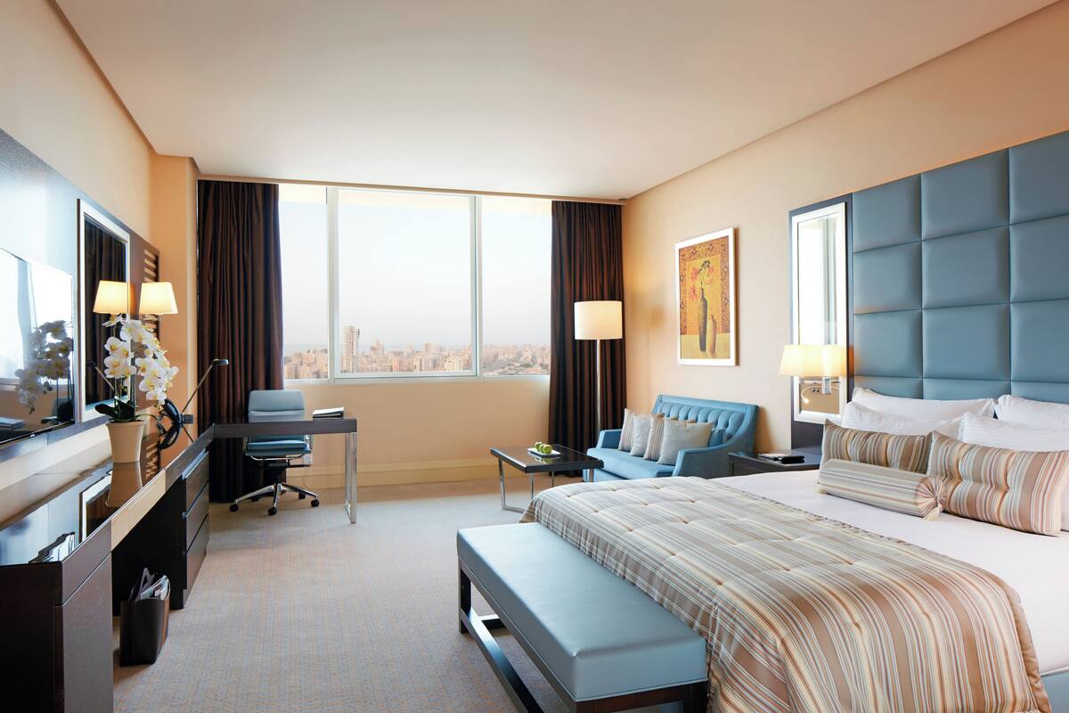 Une chambre d’hôtel avec un grand lit, un bureau avec une chaise, une petite table, un banc et une vue sur la ville grâce à de grandes fenêtres.