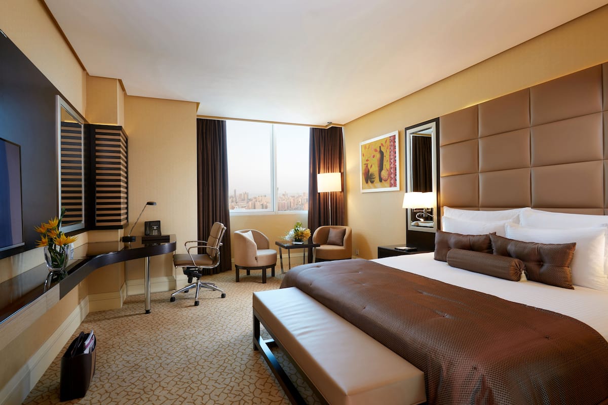 Une chambre d’hôtel avec un grand lit, un bureau avec une chaise, une petite table avec une lampe et une vue sur la ville par la fenêtre.
