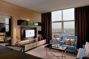 A modern hotel room with a large window, a flat-screen TV, a sofa, a coffee table, and a dining area. - Millennium Hotel and Convention Centre Kuwait (Salmiya)