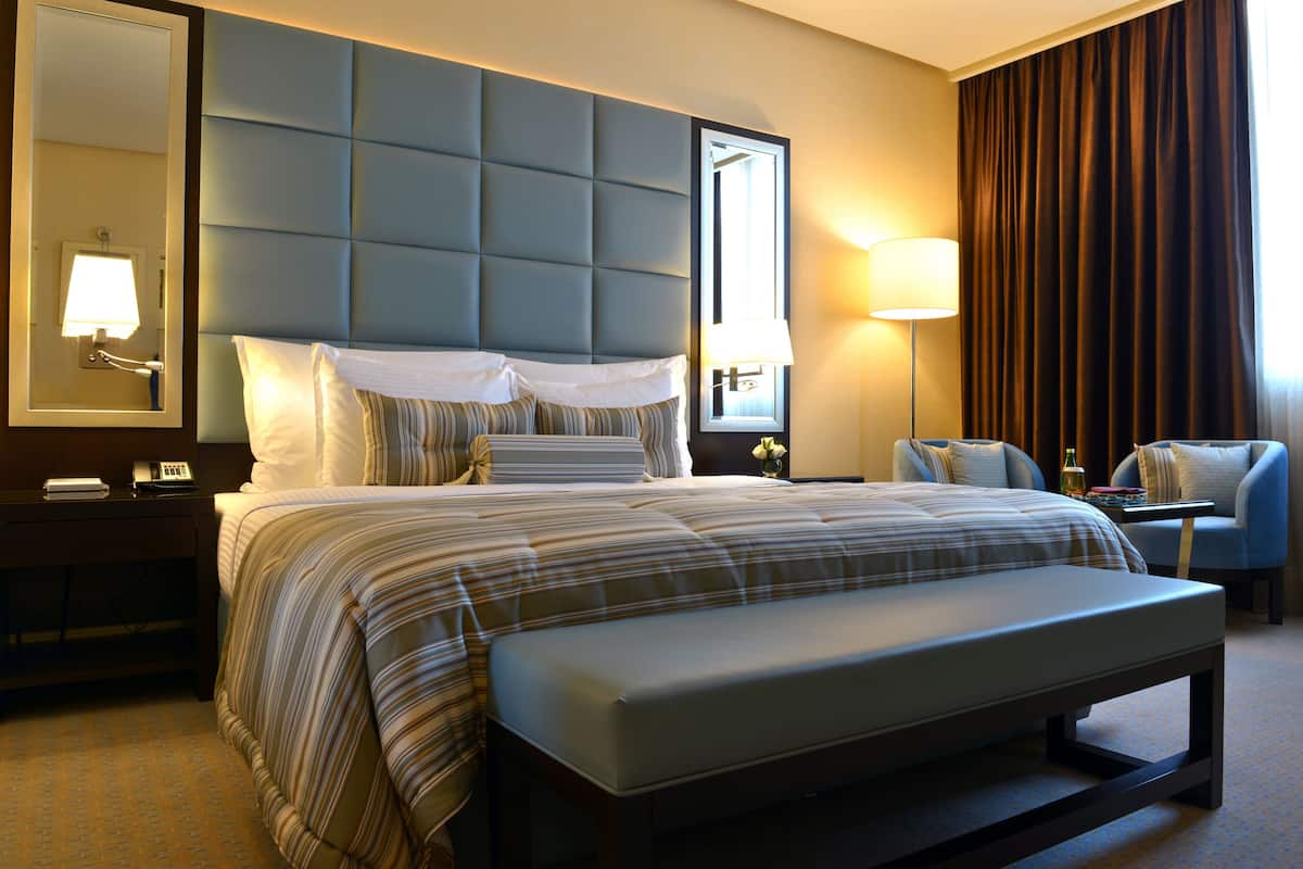 Une chambre d’hôtel avec un grand lit, une banquette, un bureau, une chaise, une table de chevet, une lampe et un miroir.