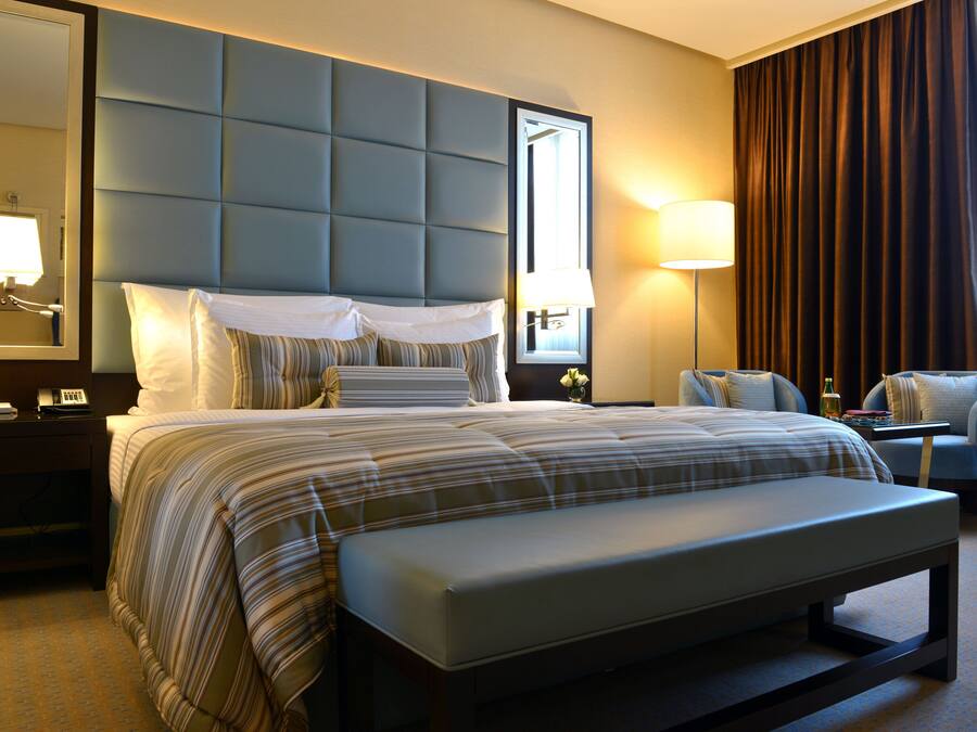 A hotel room with a large bed, a bench, a desk, a chair, a nightstand, a lamp, and a mirror.