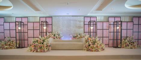 A stage with a sofa, surrounded by floral arrangements, in a room with a high ceiling and recessed lighting.