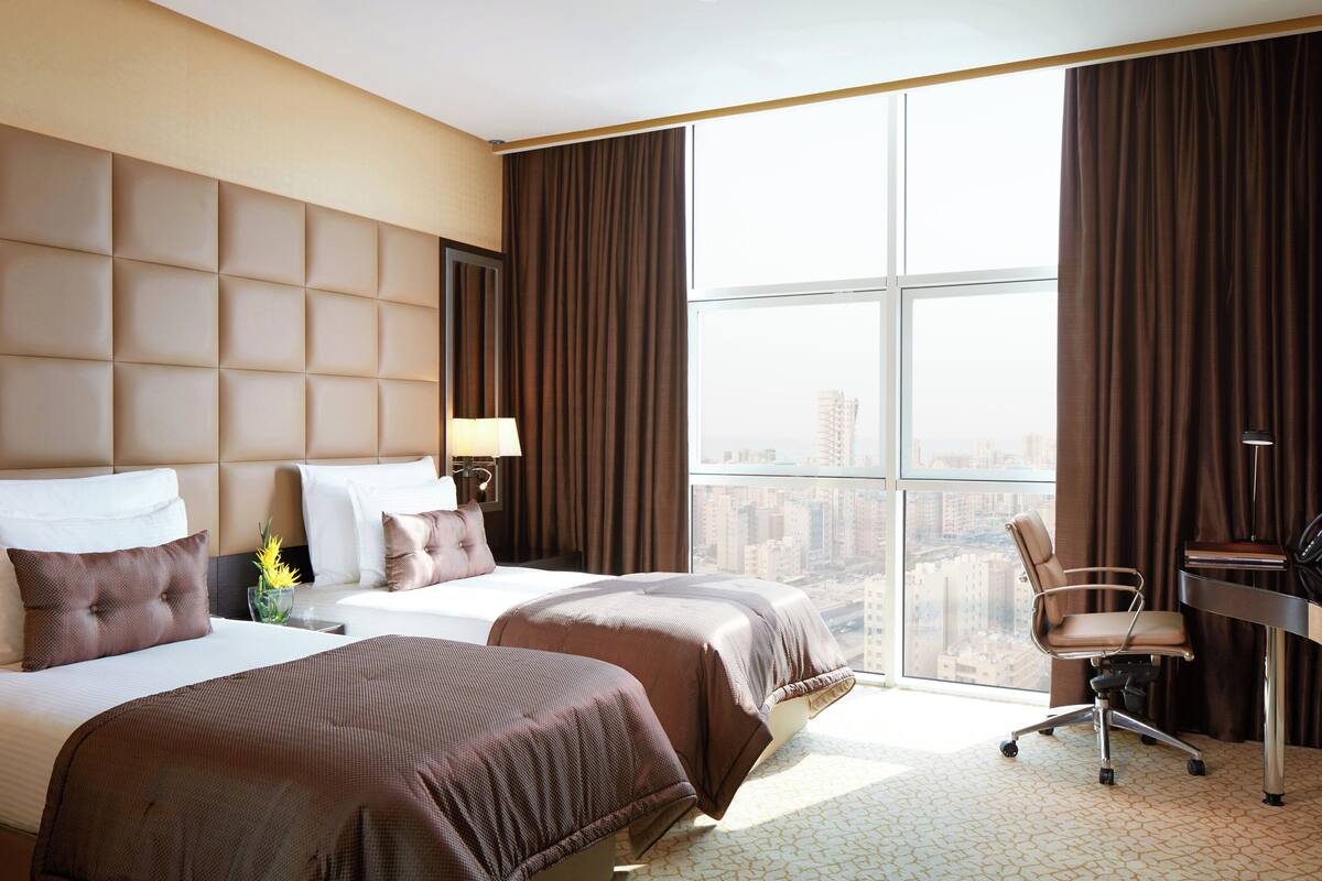 Une chambre d’hôtel avec deux lits, un bureau, une chaise et une grande fenêtre donnant sur la ville.