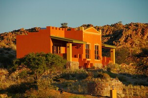 Un edificio moderno con techo plano y base de piedra, rodeado de pastizales secos y árboles dispersos.