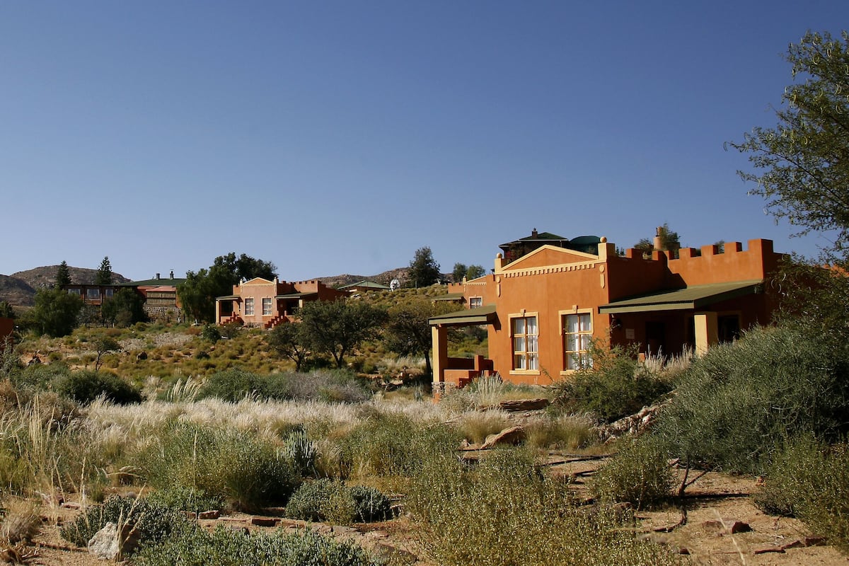 A desert landscape with a windmill, a building with a red facade, and a mountain in the background.