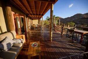 Una terraza de madera con mobiliario exterior, que incluye un sofá y mesas, con vistas a un paisaje montañoso.