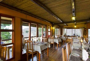 A dining area with wooden tables and chairs, large windows, and a thatched roof.