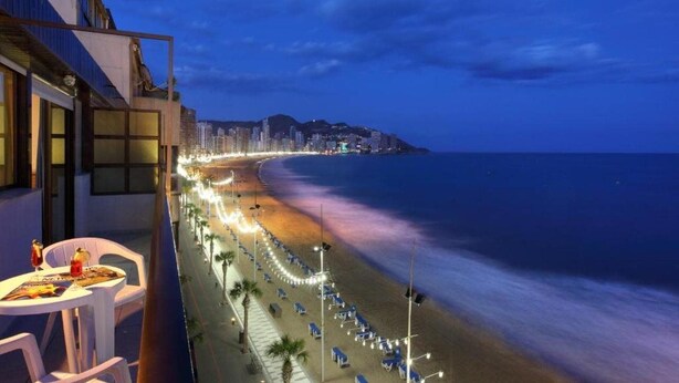 A balcony with a table and chairs overlooking a beach with rows of sun loungers and a cityscape in the background. - Bilbaino (Benidorm)