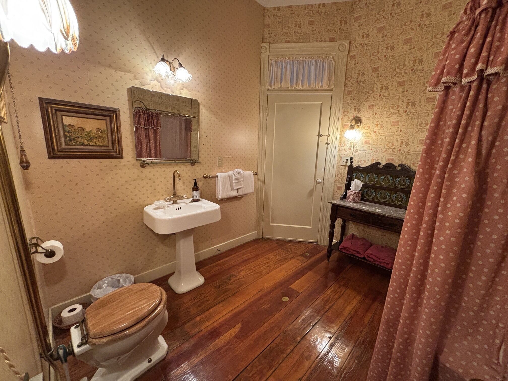 Un baño con inodoro de madera, lavamanos blanco sobre pedestal, espejo y tocador con cabecera decorativa.