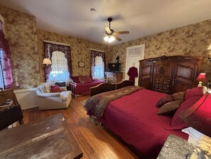 A bedroom with a wooden floor, a bed with red bedding, a wooden wardrobe, a chair, a desk, and a ceiling fan.