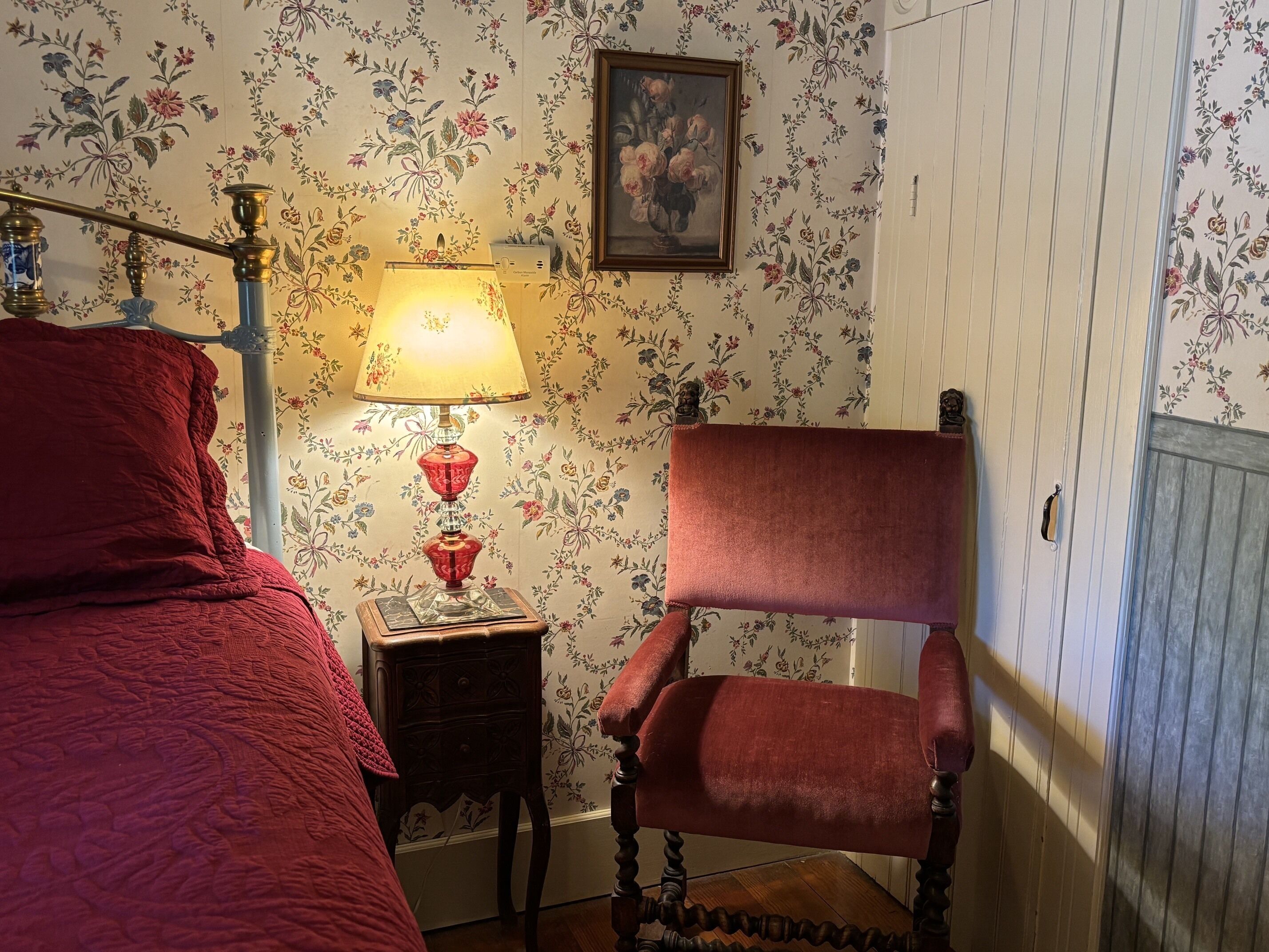 Ein Schlafzimmer mit Bett, Sessel, Nachttisch mit Lampe, floralem Wandpapier und einem Gemälde.