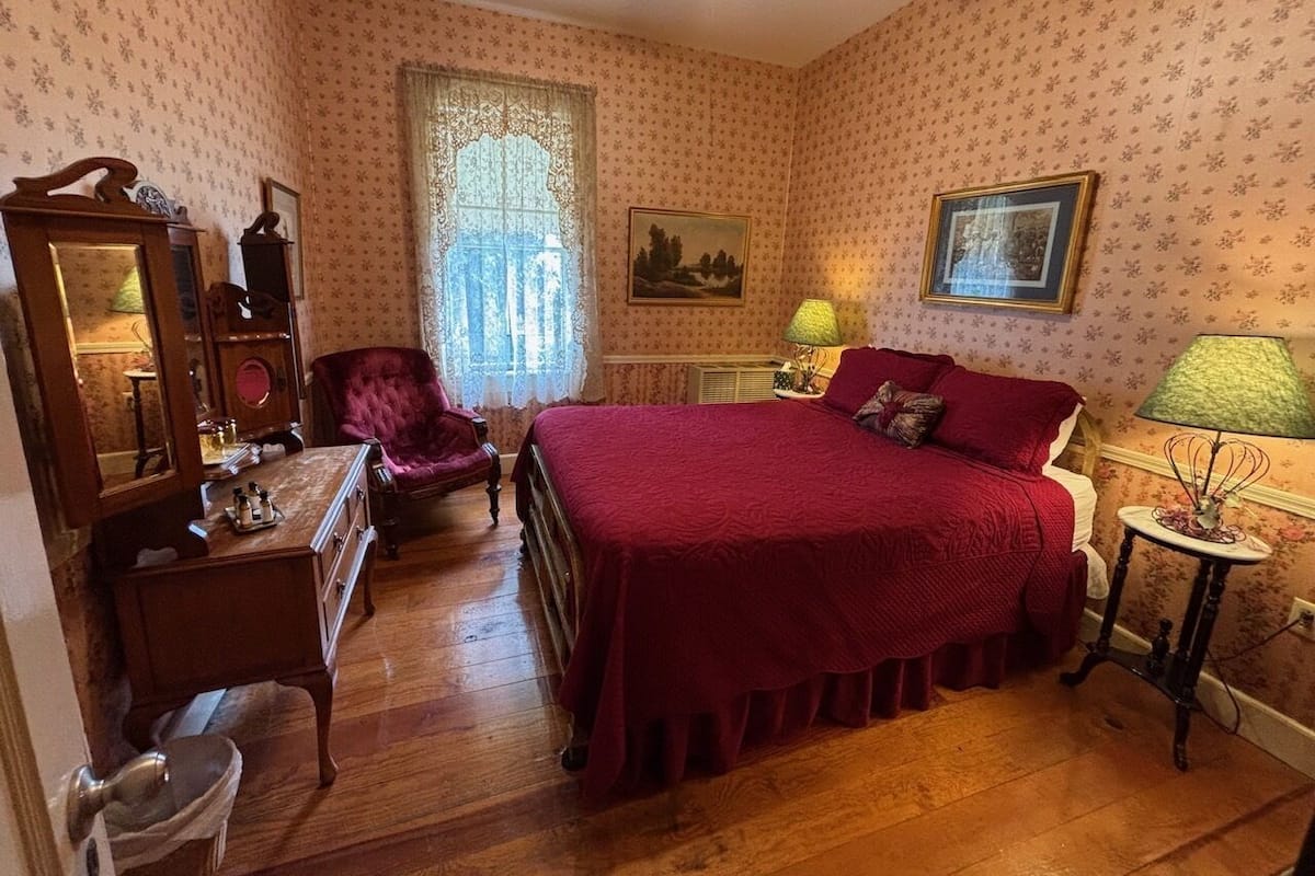 Un dormitorio con cama, tocador, cómoda, silla y ventana con cortinas de encaje.