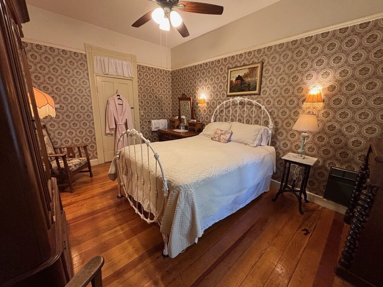 A bedroom with a white bed, wooden floor, a ceiling fan, a nightstand, a lamp, and patterned wallpaper.
