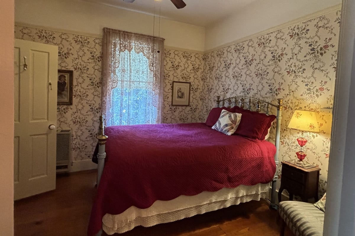 Un dormitorio con cama, ventilador de techo, ventana con cortinas de encaje y mesita de noche con lámpara.